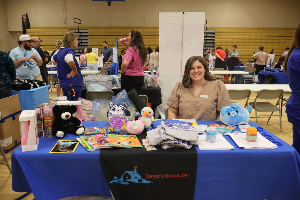 There is a woman sitting at a table that has a blue table cloth with toys sitting on top of it. There are also people in the background. 
