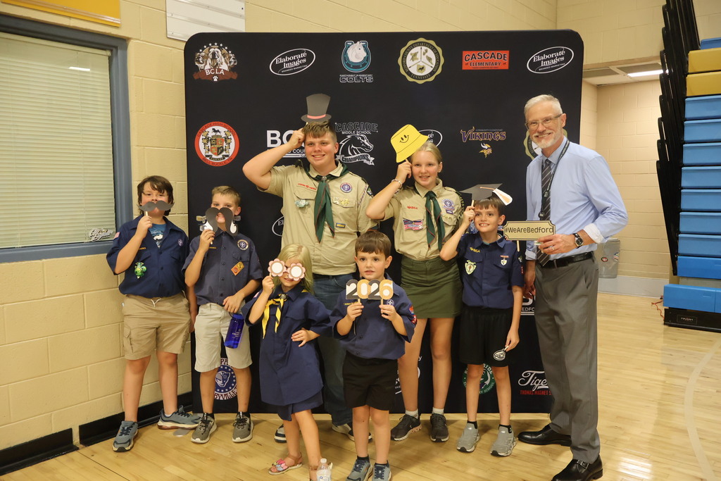 The superintendent is posed for a group picture with seven children who are all holding a funny photo prop as they all stand in front of a backdrop that has school logos.