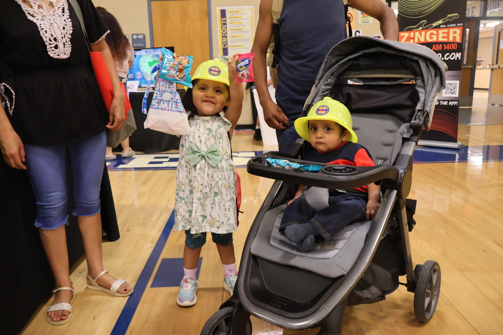 There is a baby sitting in a stroller beside a little girl who is holding up snacks while both are wearing yellow hats. 