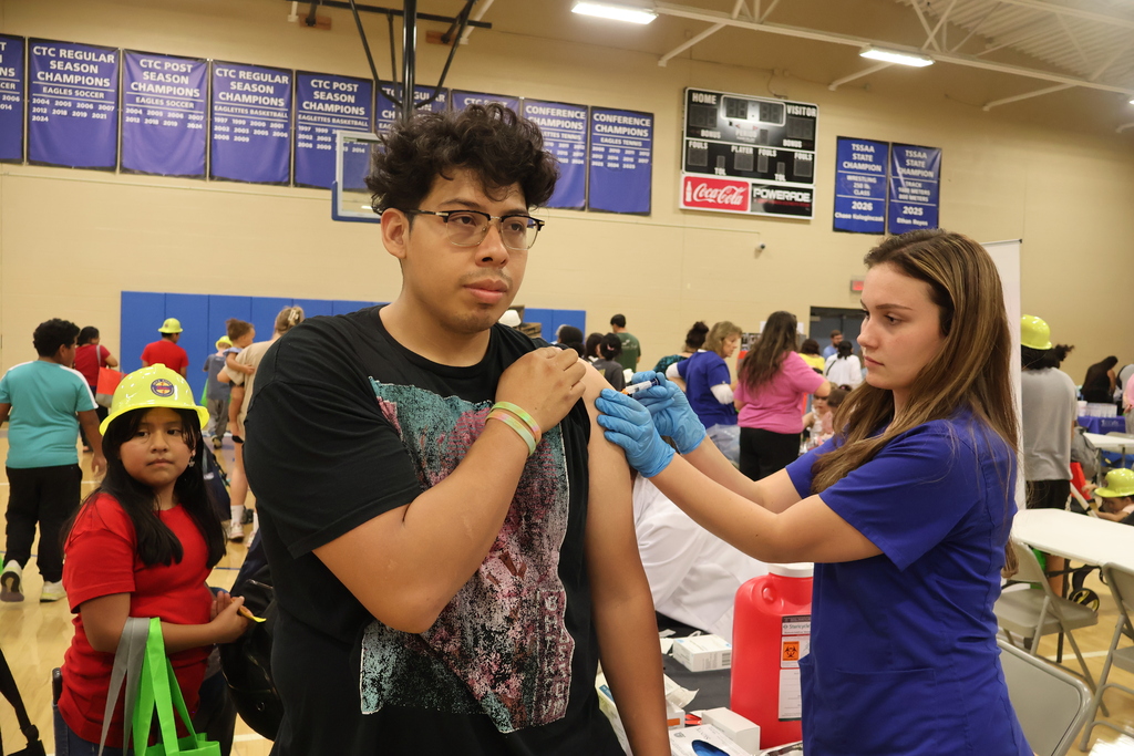 A man is holding up his shirt sleeve as he receives a shot from a woman in his arm. 