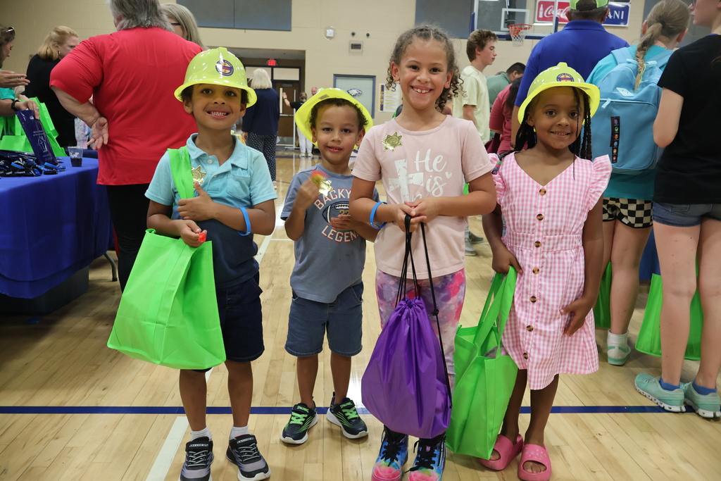 There are four children posing for a group picture in the gym of a school with some holding green and purple bags and wearing yellow hats. 