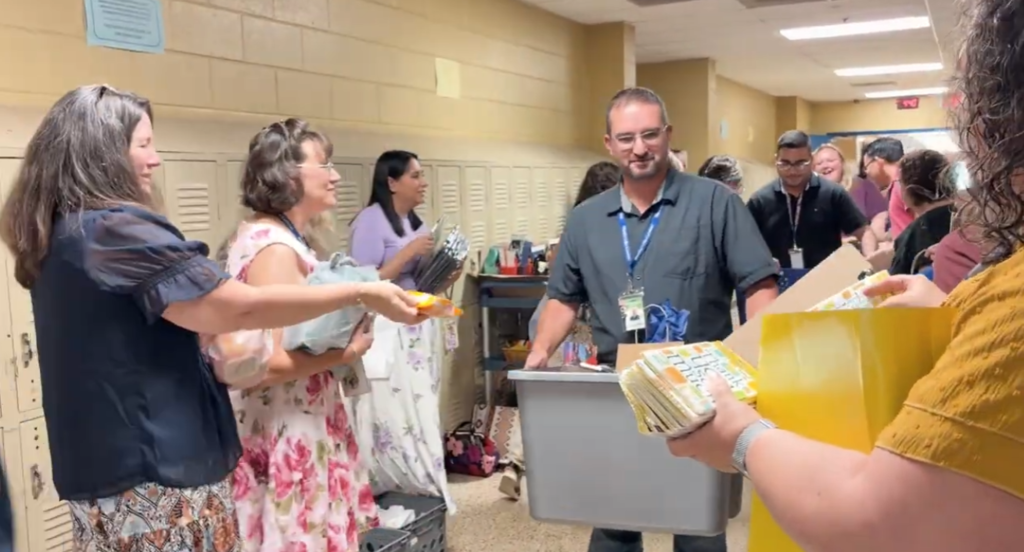 This is a picture of a group of teachers in the hallway all handing school supplies to a new teacher who is holding a gray tote.