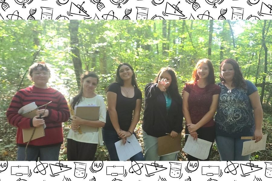 Six freshman Physical Science students holding clipboards and smiling in wooded forest setting. 