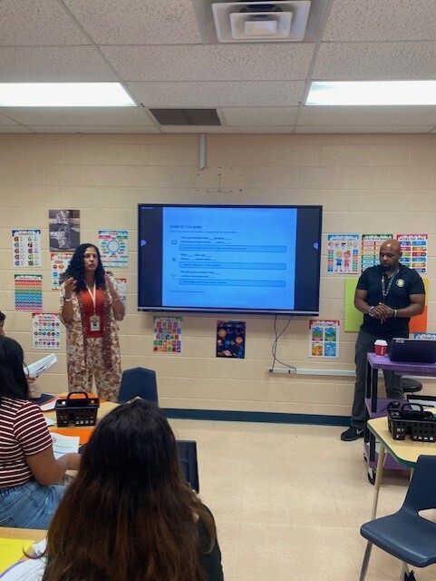 There are two people in this picture who are standing beside a large screen in a classroom giving a presentation to families who are attending.
