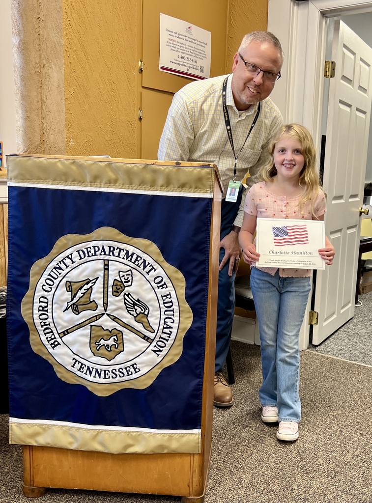This is a picture of a little girl who is holding up a certificate for starting the pledge of allegiance at the school board meeting and she is standing beside a podium as well as the assistant superintendent.