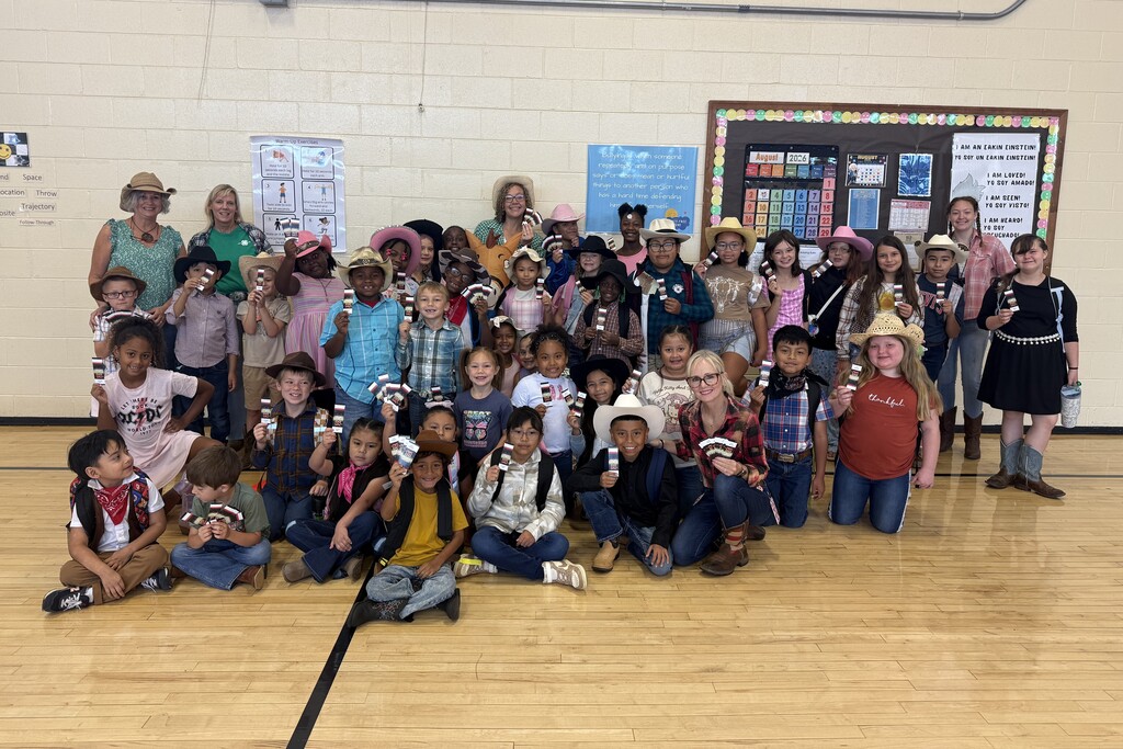 There is a large group of students and a few teachers all posed for a picture in their gym while wearing cowboy apparel and holding up tickets to the Celebration.