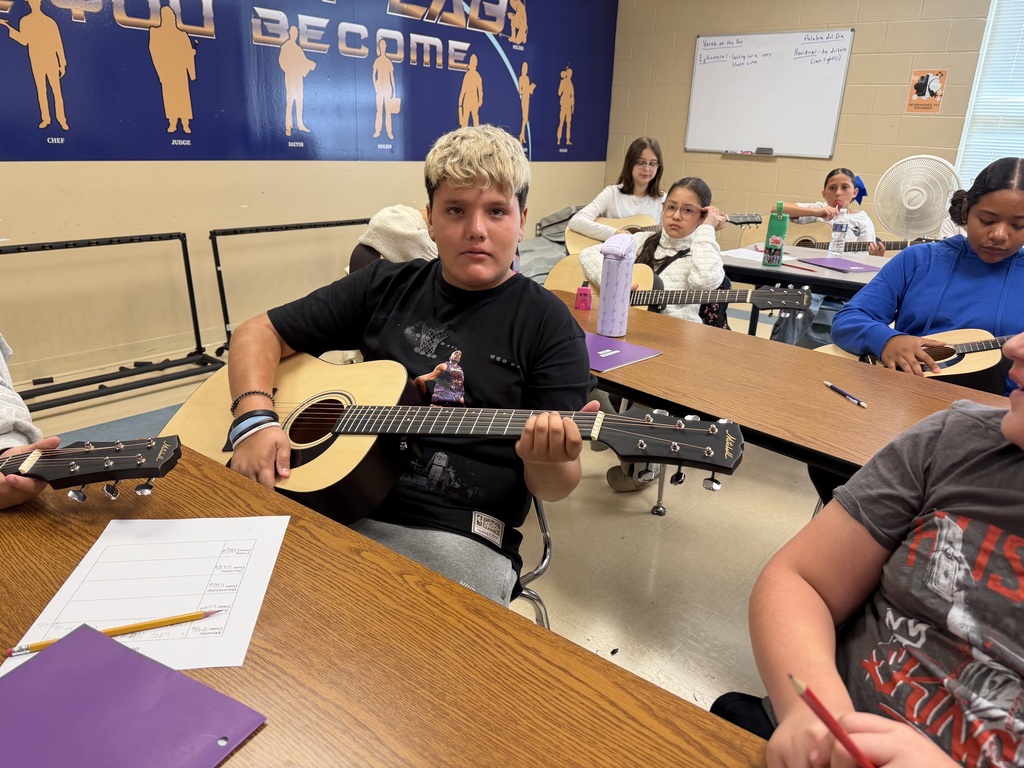 There are several students sitting at a desk in a classroom and each are holding a guitar.