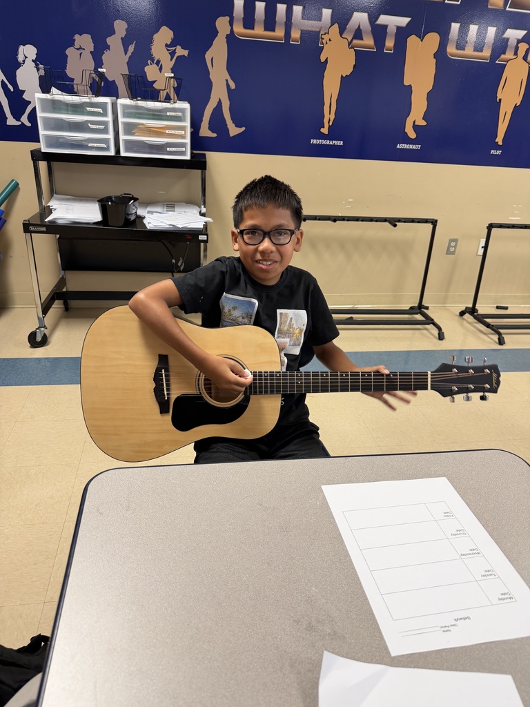 A student is sitting at a table in a classroom and he is holding a guitar.