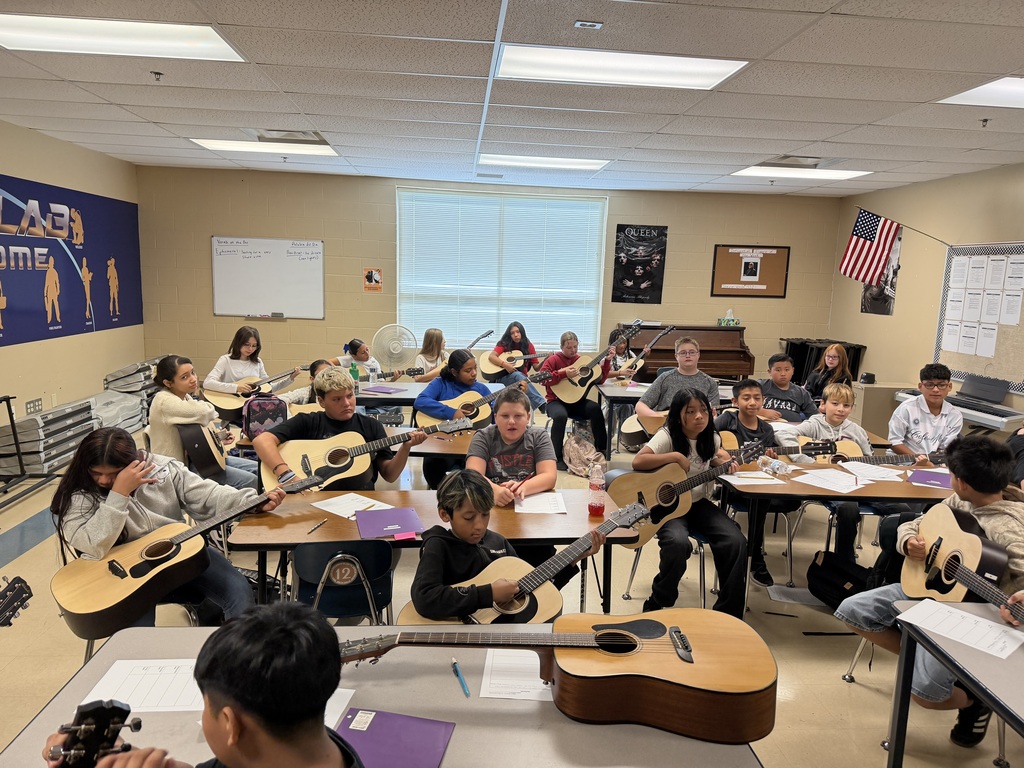 There is a group of students all sitting at tables in their classroom and each are holding a guitar that was received through a teacher grant.