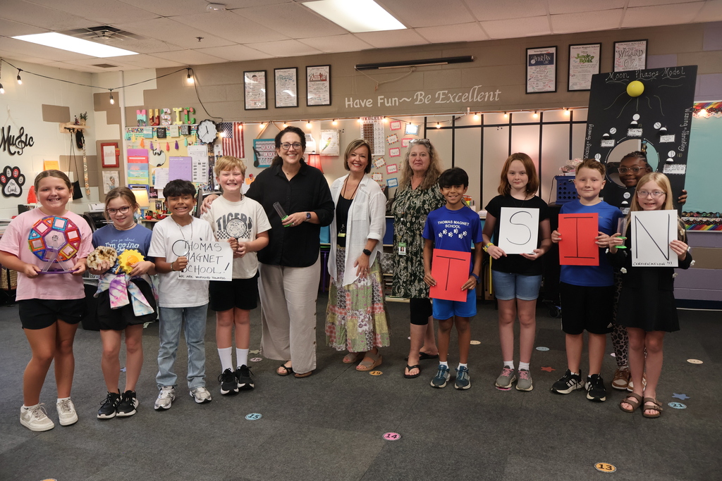 There are three teachers and nine students all posed for a group picture in the music classroom while they are all holding up science related items used in the classroom. 