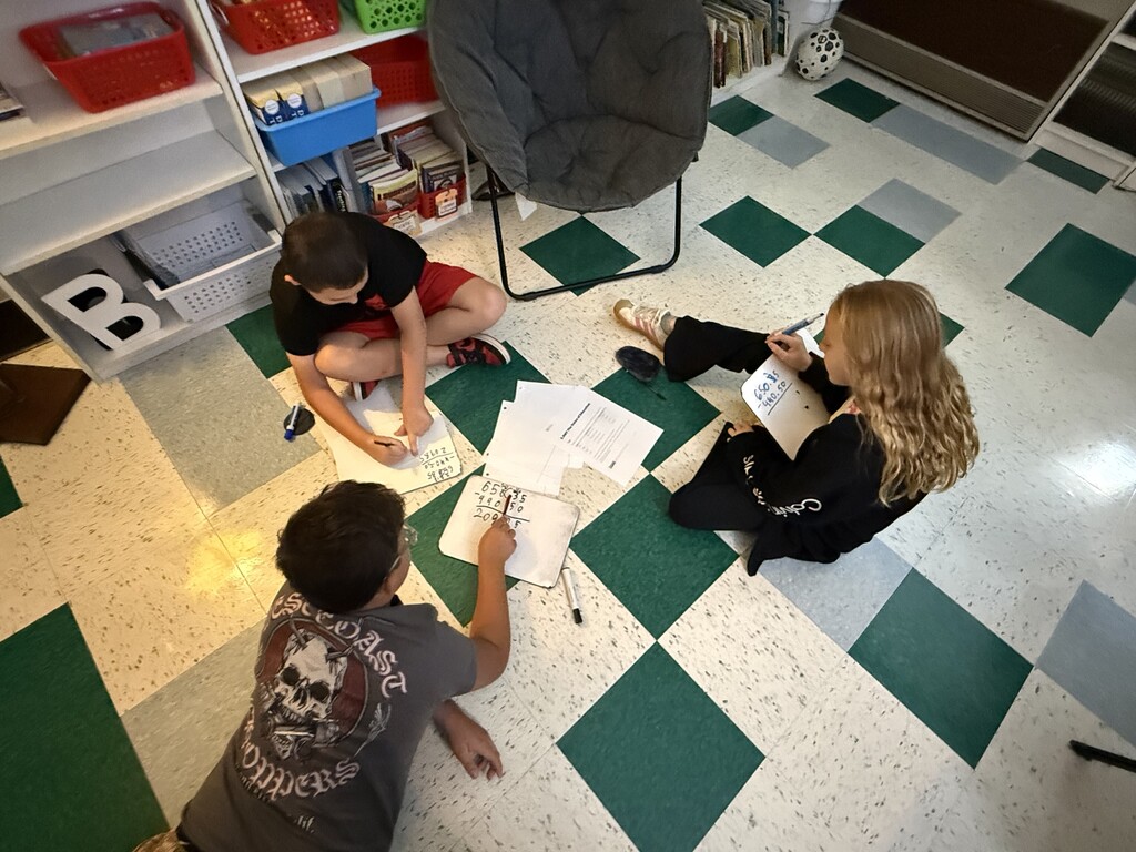 There are three students sitting on the floor all working on math problems in their classroom.
