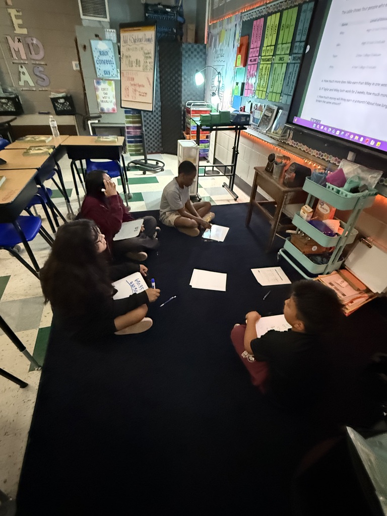 There are several students sitting on a black carpet all working on math problems in their classroom.
