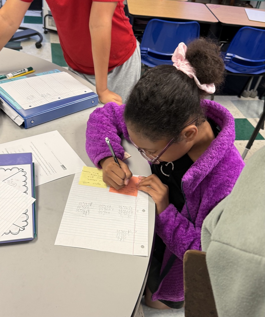 A student wearing a purple jacket is sitting at a table while working on math problems in the classroom.