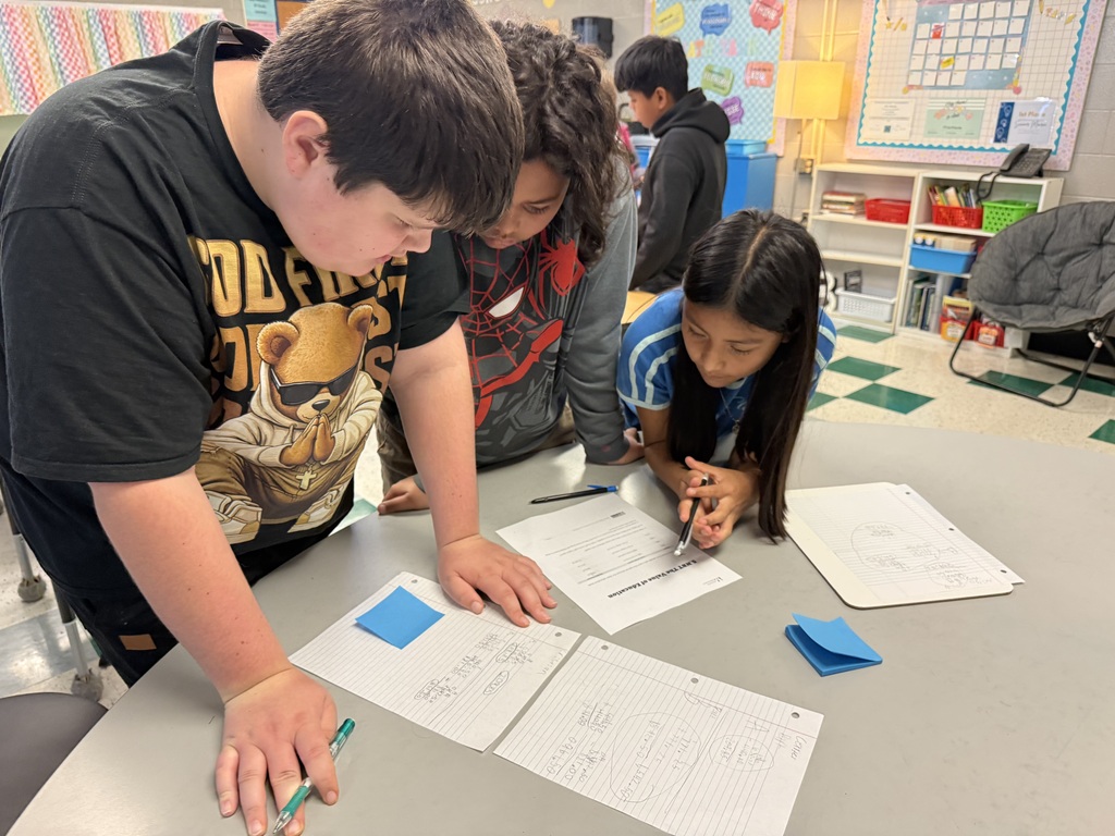 There are three students standing at a table while they are all working on math problems in their classroom.
