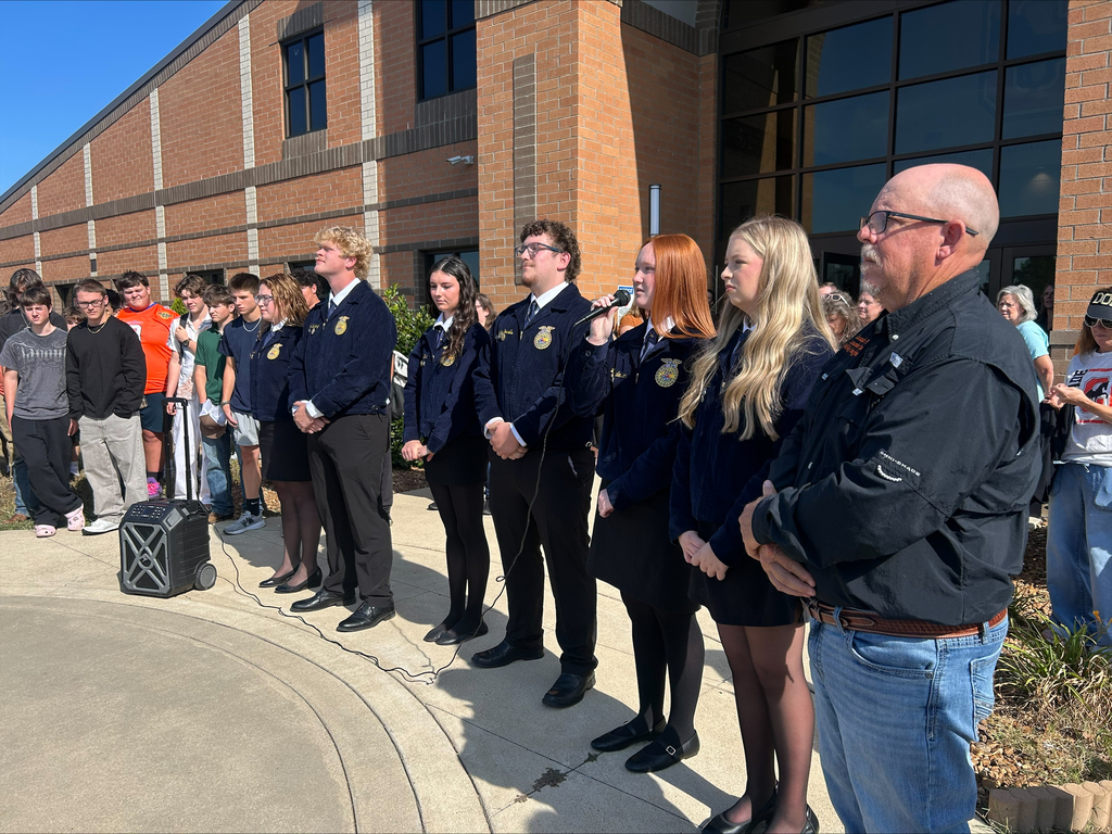 FFA students conducting flag raising ceremony outside school building, with student speaking into microphone while others observe.