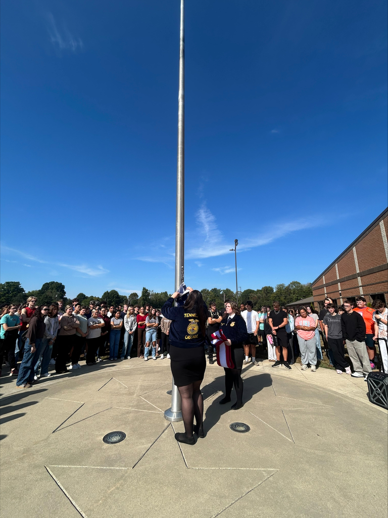 Large group of students and staff gathered around flagpole during 9/11 memorial flag raising ceremony on a sunny day.