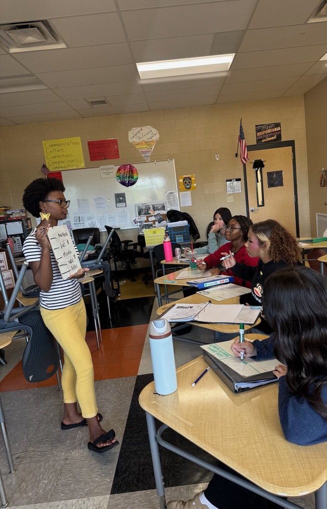 National Honor Society student tutoring Spanish I students during Champ Time, standing at front of classroom holding presentation sign while peers take notes at desks.