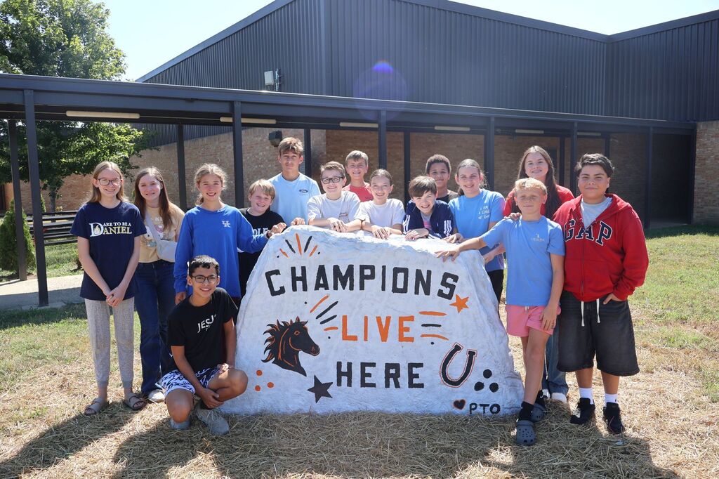 There is a group of students standing outside by their school with the newly donated spirit rock that is painted "Champions Live Here".
