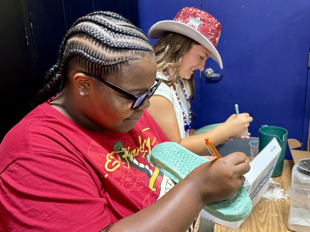 Two students are in a classroom sitting at a table making a cast of a shoe that they are holding. 