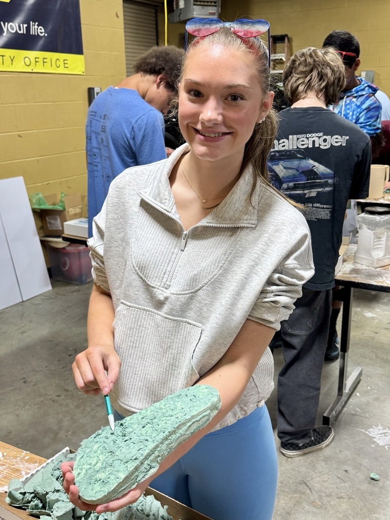 A student is standing in a classroom at a table making a cast of a shoe that she is holding. 
