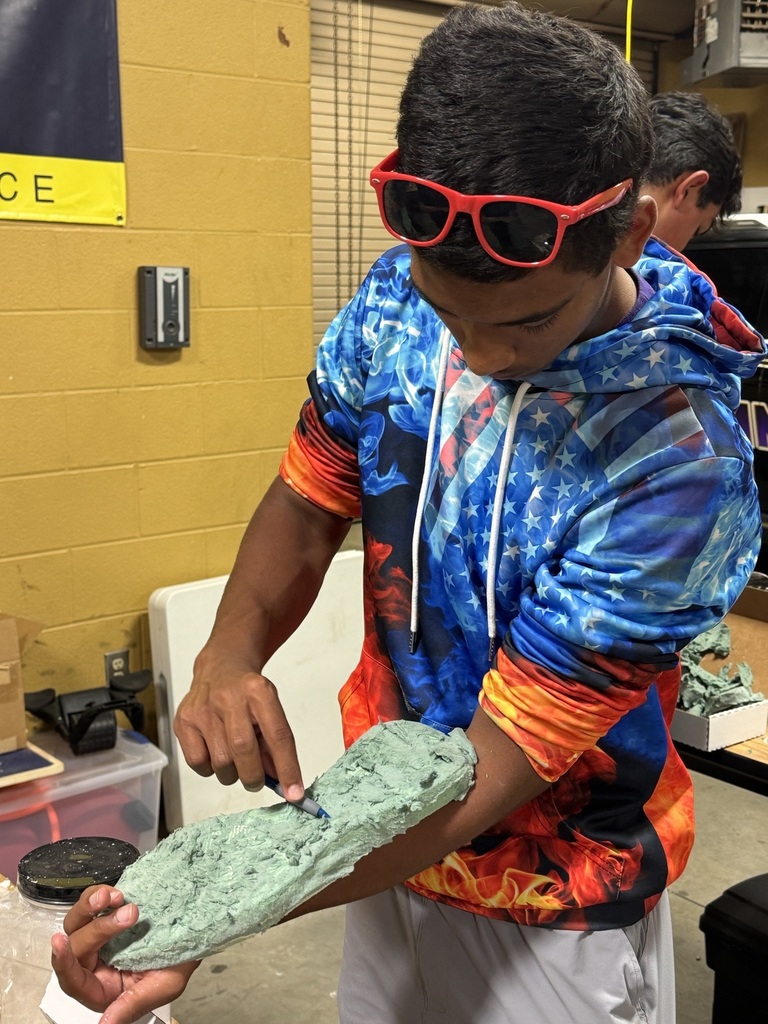 A student is standing in a classroom at a table making a cast of a shoe that he is holding. 
