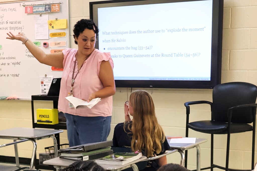A teacher is standing before her class reading a story to them as they read along from the same book. 