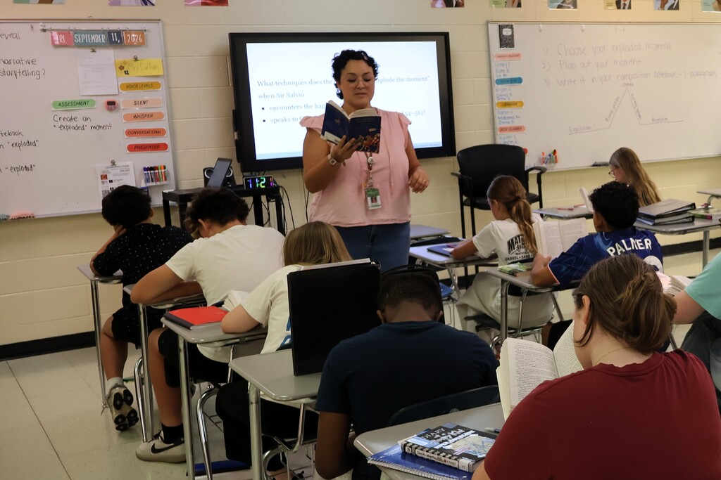 A teacher is standing before her class reading a story to them as they read along from the same book. 