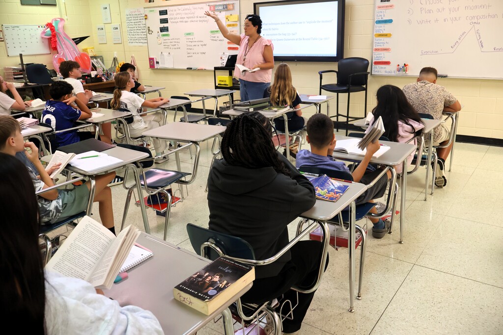 A teacher is standing before her class reading a story to them as they read along from the same book. 