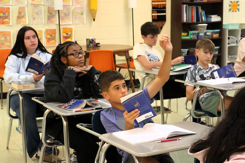 There are several students all sitting at their desks, and one student is holding his hand up to ask a question. 