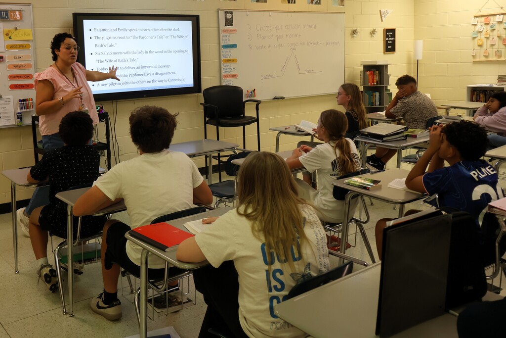 A teacher is standing before her class reading a story to them as they read along from the same book. 