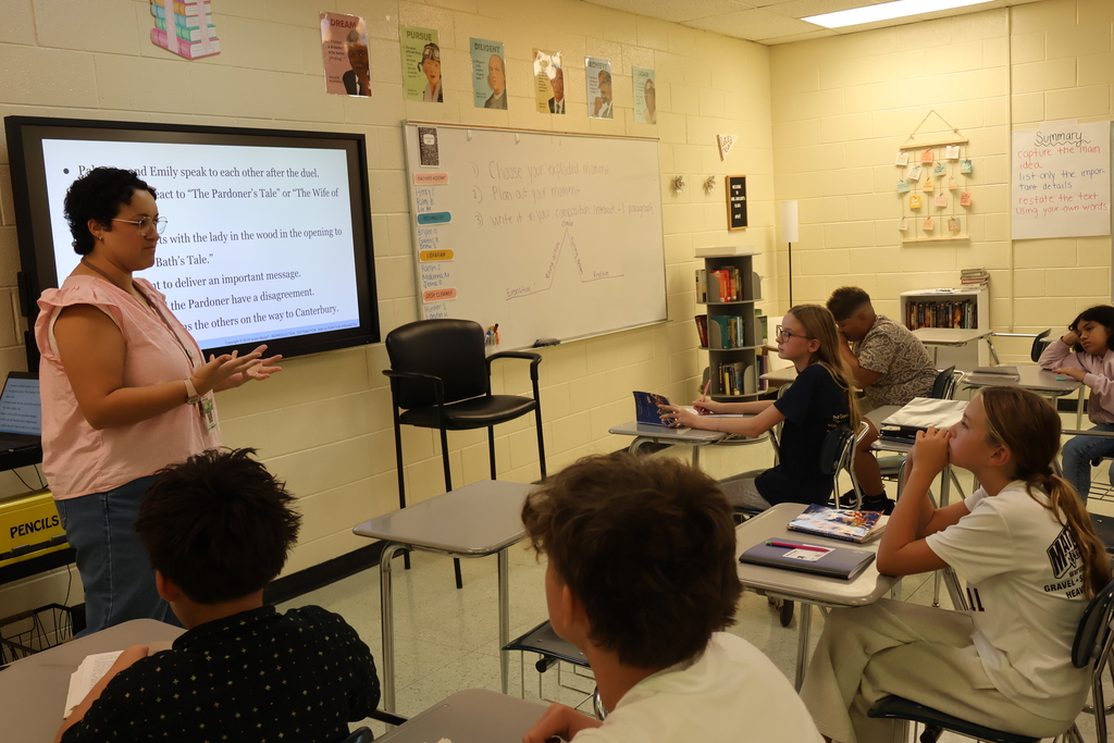 A teacher is standing before her class reading a story to them as they read along from the same book. 