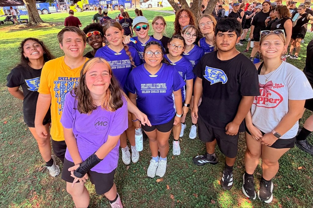 There are several students all posed for a group picture outside under a tree with other people in the background.