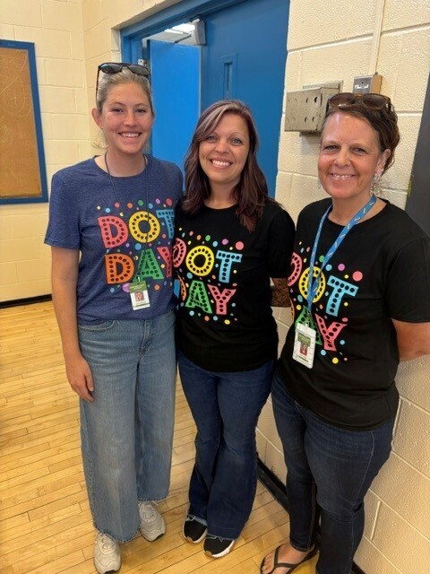 Three teachers are posed for a group picture in the gym while wearing their "Dot Day" matching shirts. 