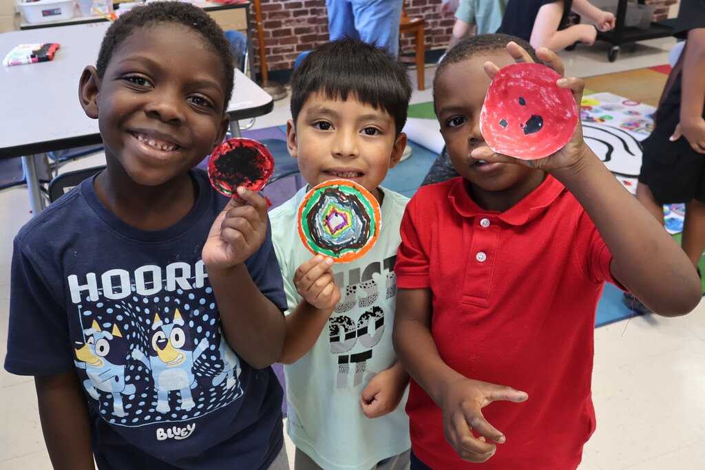 Three students are all posed for a group picture in the art classroom while holding up their dots they decorated. 