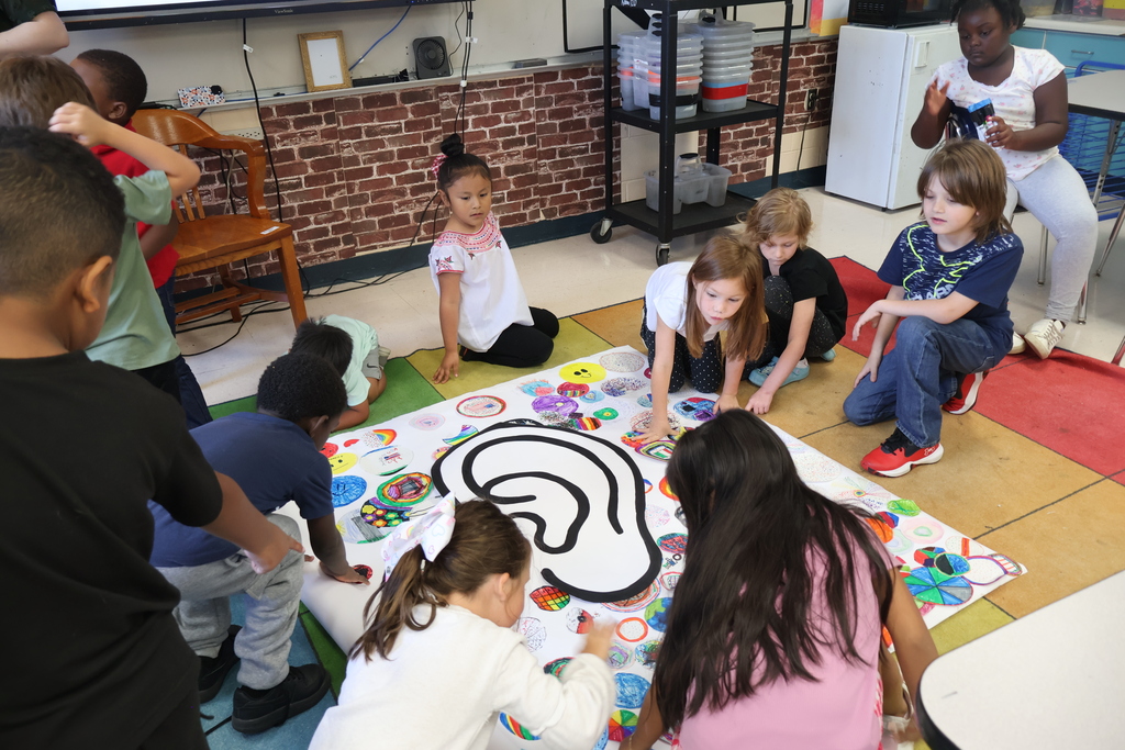 There are several students who are all sitting on the ground adding the dots they decorated to a large piece of paper that has an ear on it. 