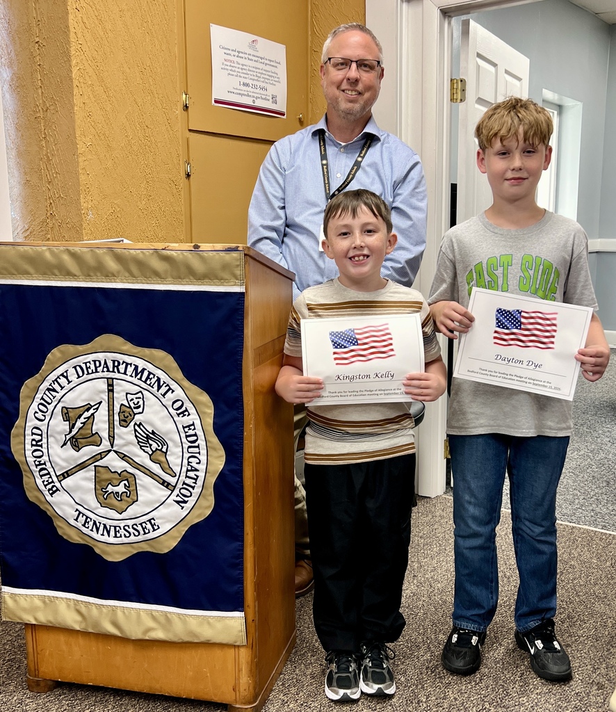 There are two students standing in the picture beside a podium with each holding a certificate. The assistant superintendent is standing behind them.