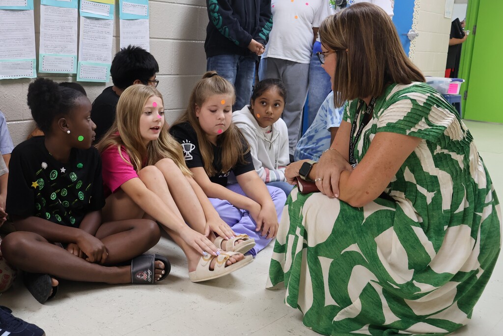 A woman in a green dress is kneeling down in front of students who are sitting on the floor as they discuss their work with her that is hanging on the wall. 