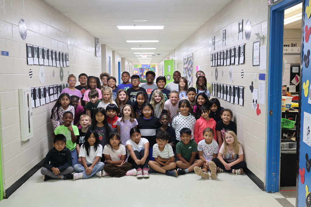 All of the third graders are in the hallway posing for a picture with their black and white pictures hanging on the walls beside them. 