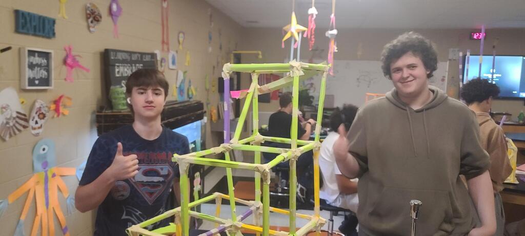 Physics students working in groups building roller coaster models from colorful craft sticks, straws, and tape in classroom after learning about linear velocity, tangential velocity, and centripetal acceleration.