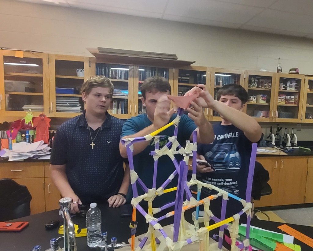 Physics students working in groups building roller coaster models from colorful craft sticks, straws, and tape in classroom after learning about linear velocity, tangential velocity, and centripetal acceleration.