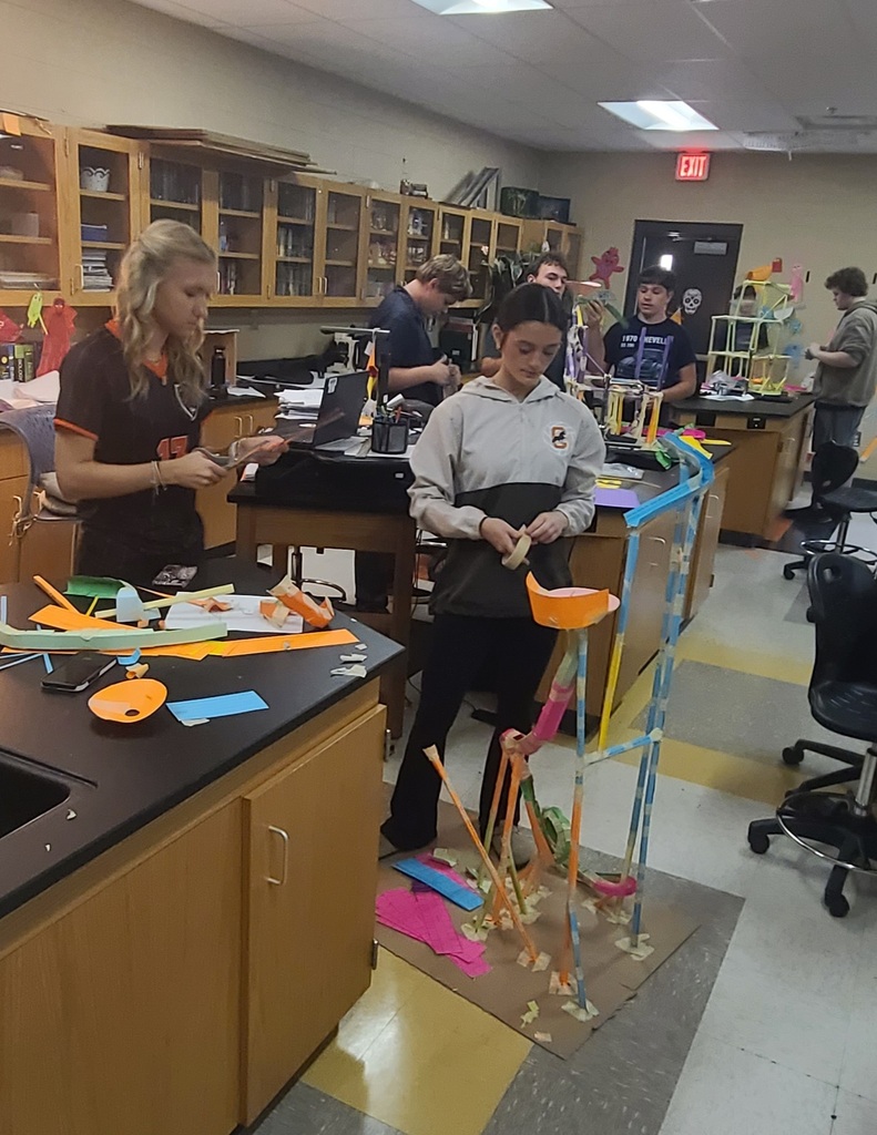 Physics students working in groups building roller coaster models from colorful craft sticks, straws, and tape in classroom after learning about linear velocity, tangential velocity, and centripetal acceleration.