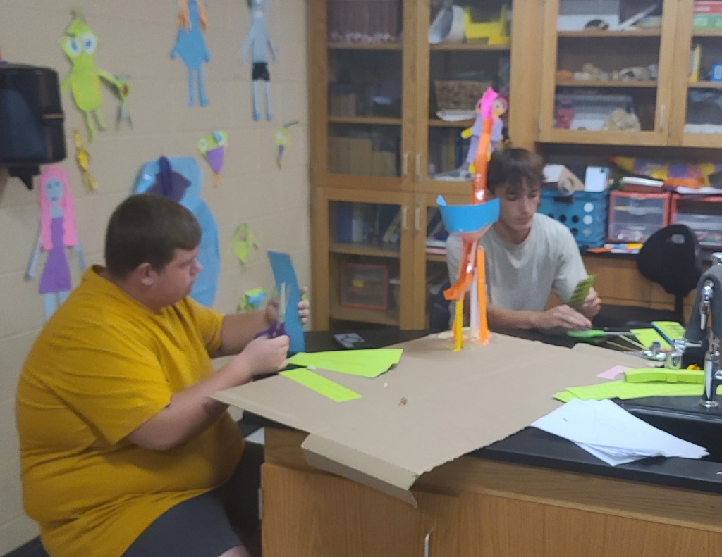 Physics students working in groups building roller coaster models from colorful craft sticks, straws, and tape in classroom after learning about linear velocity, tangential velocity, and centripetal acceleration.