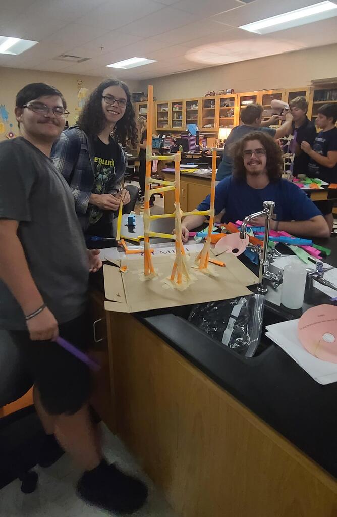Physics students working in groups building roller coaster models from colorful craft sticks, straws, and tape in classroom after learning about linear velocity, tangential velocity, and centripetal acceleration.