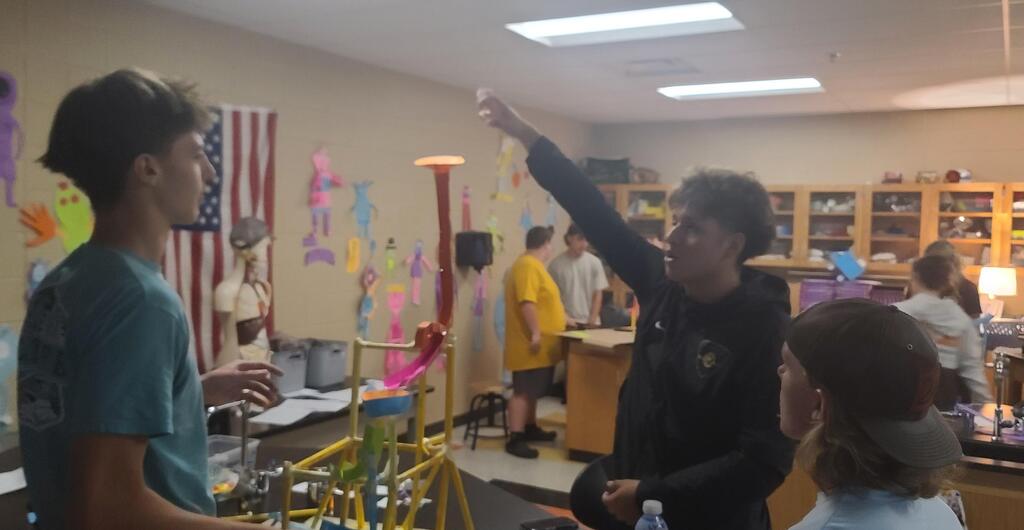 Physics students working in groups building roller coaster models from colorful craft sticks, straws, and tape in classroom after learning about linear velocity, tangential velocity, and centripetal acceleration.