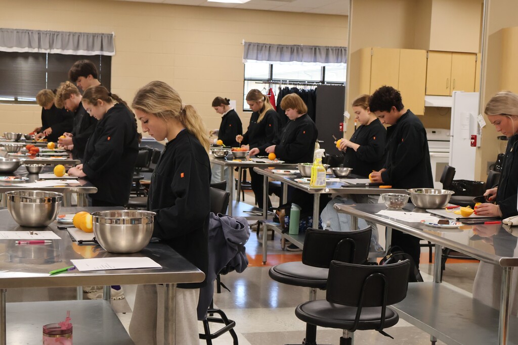 There are several students all standing at their silver tables with bowls of fruits and vegetables as they are learning to cut orange peels to use as a garnish. 
