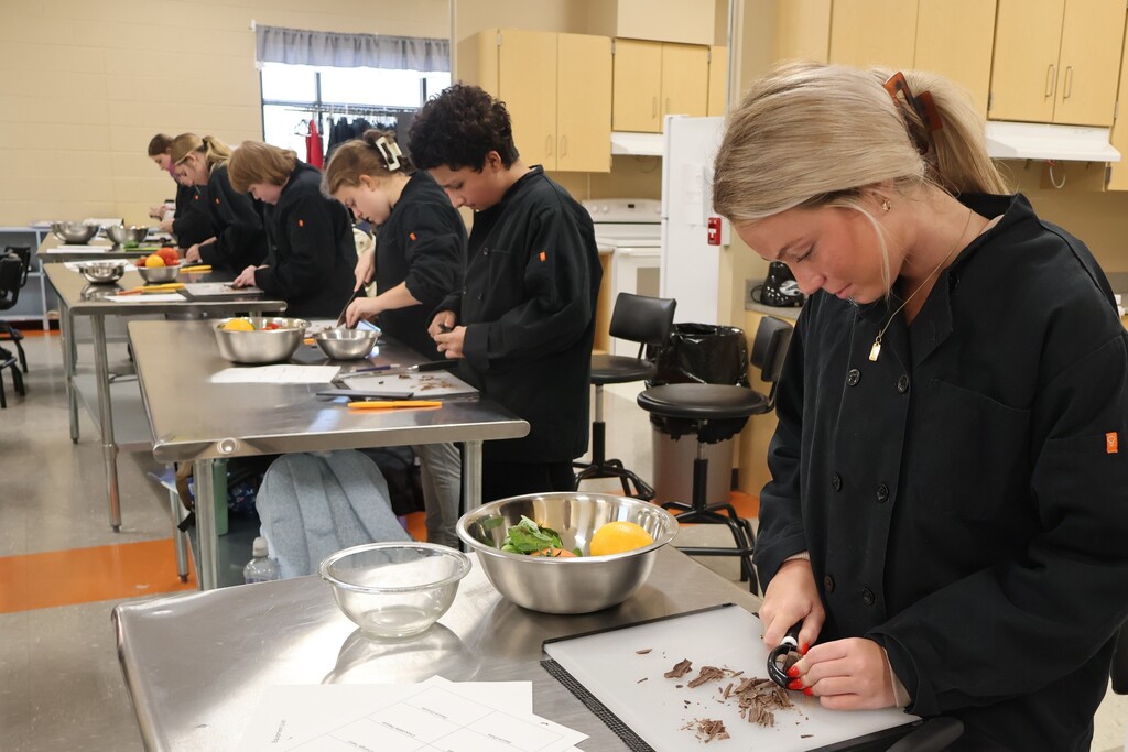 There are several students all standing at their silver tables with bowls of fruits and vegetables and they are learning to cut chocolate as a garnish. 