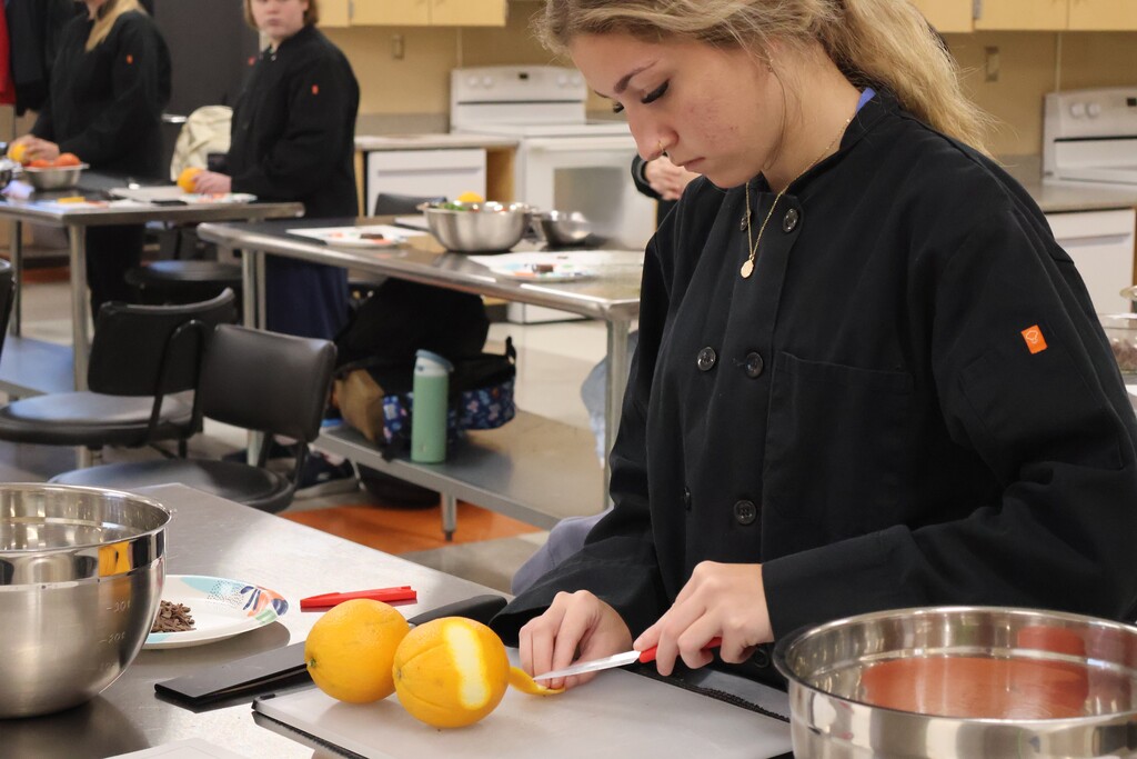 A student is standing at a silver table as she is practicing cutting an orange peel to use as a garnish. 