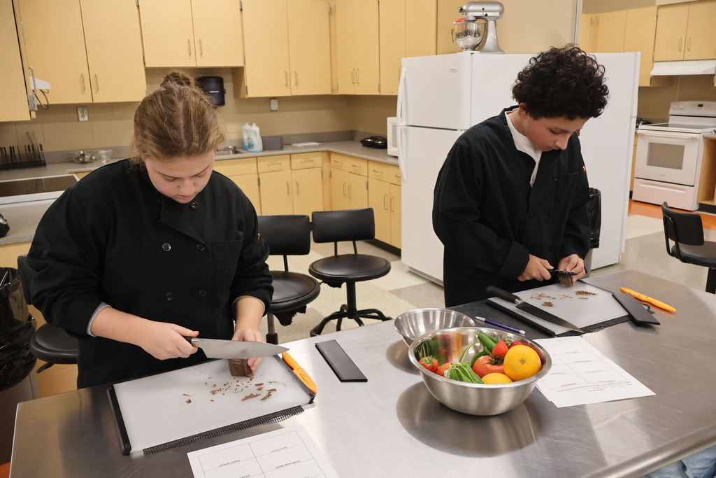 There are two students standing at their silver table with bowls of fruits and vegetables as they are learning to cut chocolate as a garnish. 