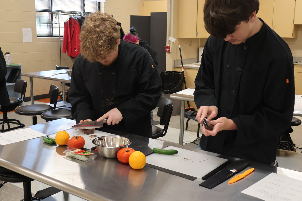 There are two students standing at their silver table with bowls of fruits and vegetables as they are learning to cut chocolate as a garnish. 
