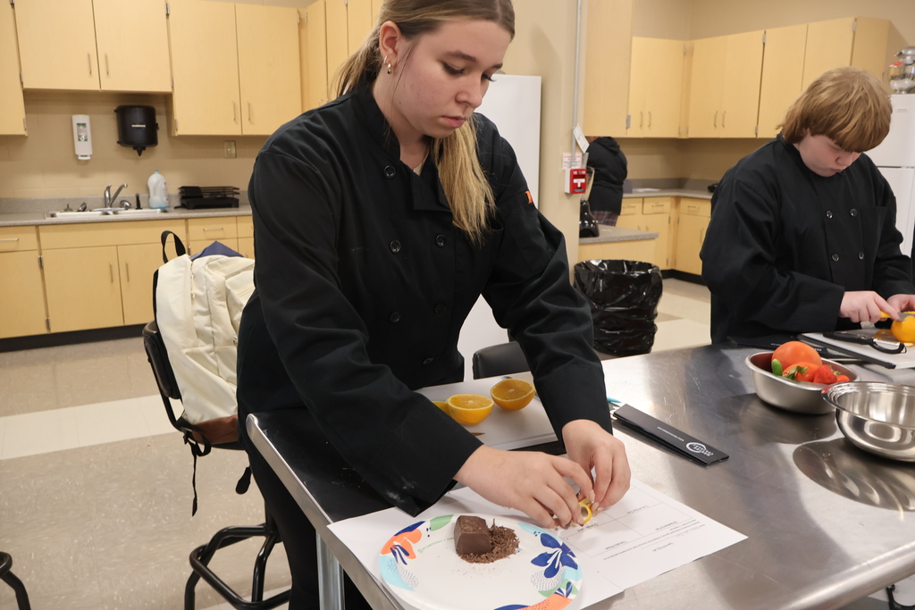 A student is standing at a silver table as she is practicing cutting an orange peel to use as a garnish. 